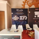 iFlair booth at Laracon India 2025 with a dark backdrop, white chairs and red carpet